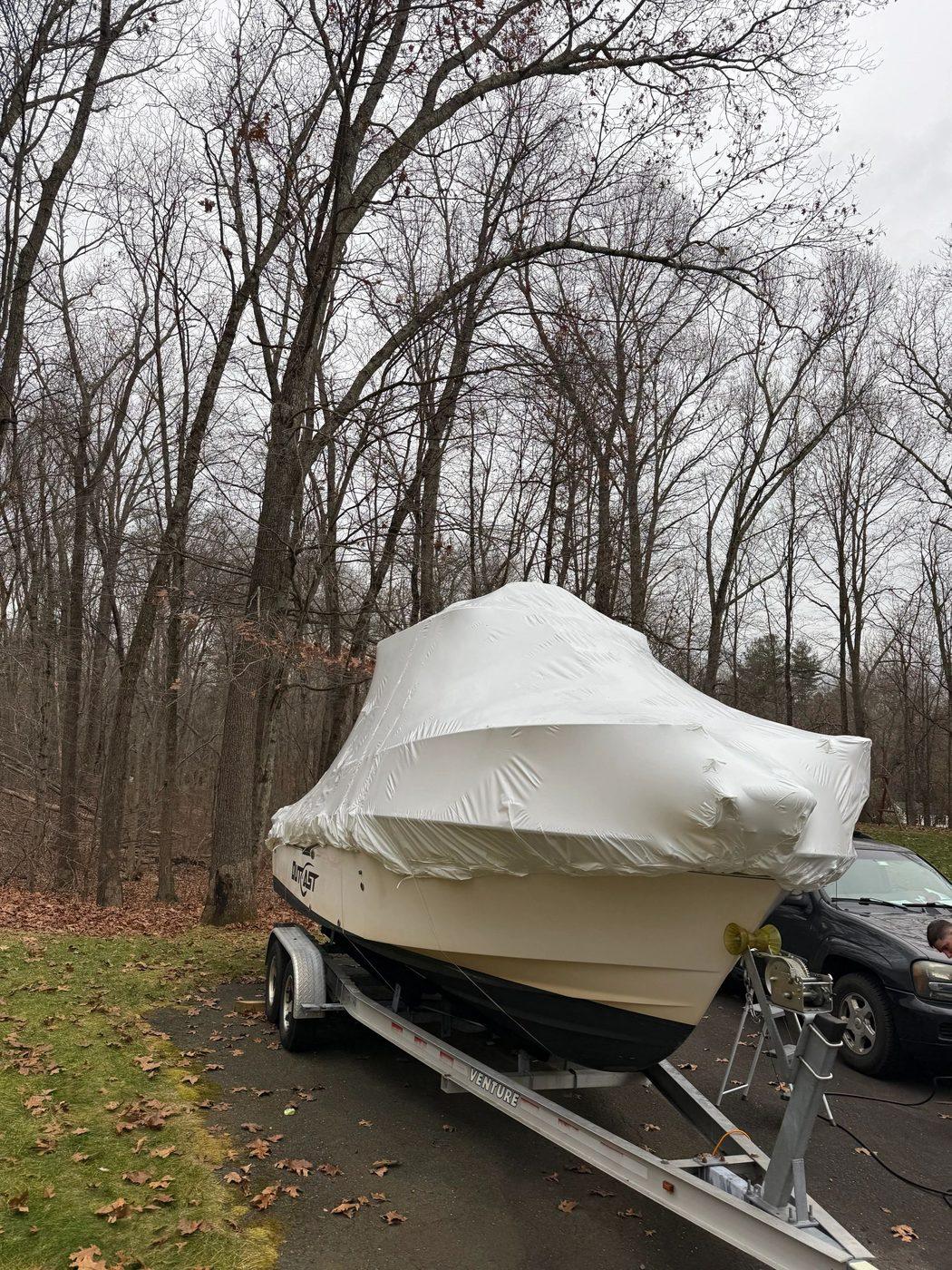 Boat covered with a white tarp on a trailer in a wooded lot